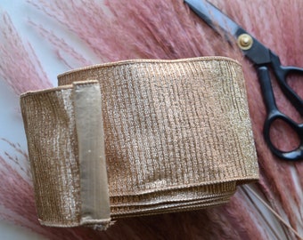 May include: A roll of gold metallic ribbon with a textured surface. The ribbon is rolled up and has a seam down the side. The ribbon is lying on a white surface.
