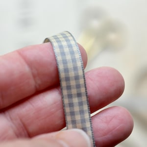 May include: A narrow ribbon featuring a repeating gingham check pattern in shades of light grey and cream. The ribbon has a smooth texture and finished edges, displaying a consistent grid design throughout its length for decorative use.
