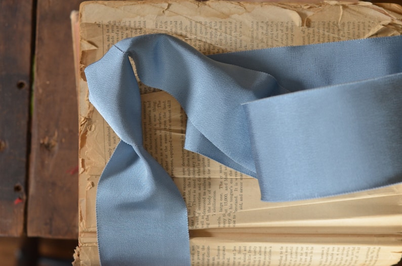 May include: A light blue ribbon is folded and placed on top of an open book. The ribbon is made of a soft, textured fabric.