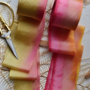May include: Two ombre silk ribbons, one yellow to pink and one pink to orange, are arranged on a white surface with dried ferns and grasses.