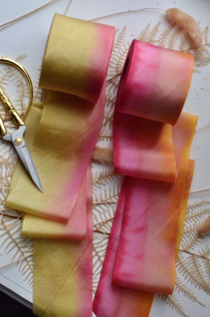 May include: Two ombre silk ribbons, one yellow to pink and one pink to orange, are arranged on a white surface with dried ferns and grasses.