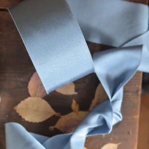 May include: A light blue satin ribbon is curled and twisted on a wooden surface with dried leaves. The ribbon is a soft, smooth fabric with a subtle sheen.