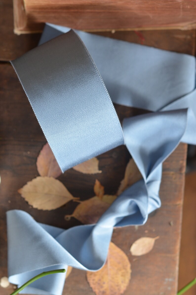 May include: A light blue satin ribbon is curled and twisted on a wooden surface with dried leaves. The ribbon is a soft, smooth fabric with a subtle sheen.