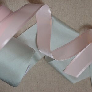 May include: Two rolls of ribbon, one light pink and one light green, are arranged on a beige fabric background.