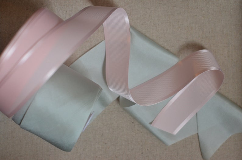 May include: Two rolls of ribbon, one light pink and one light green, are arranged on a beige fabric background.