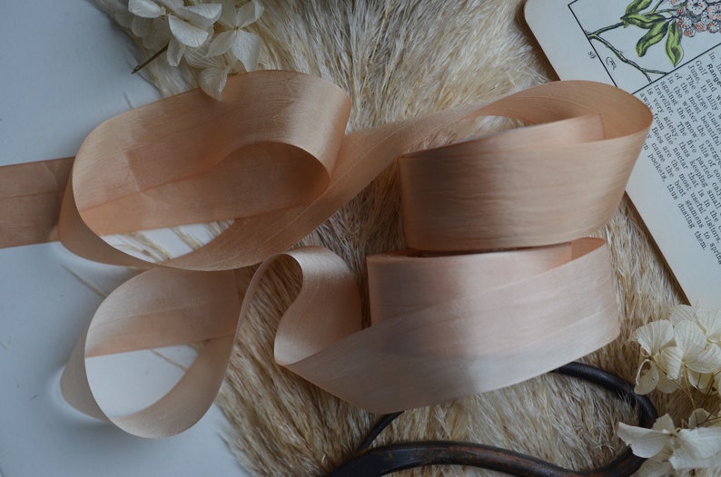 May include: A close-up of a spool of peach-colored satin ribbon. The ribbon is soft and silky, with a subtle sheen. It is perfect for adding a touch of elegance to any project.