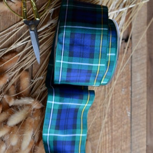 May include: A roll of blue, green, and yellow plaid ribbon with a white background. The ribbon is on a wooden surface with dried grass.