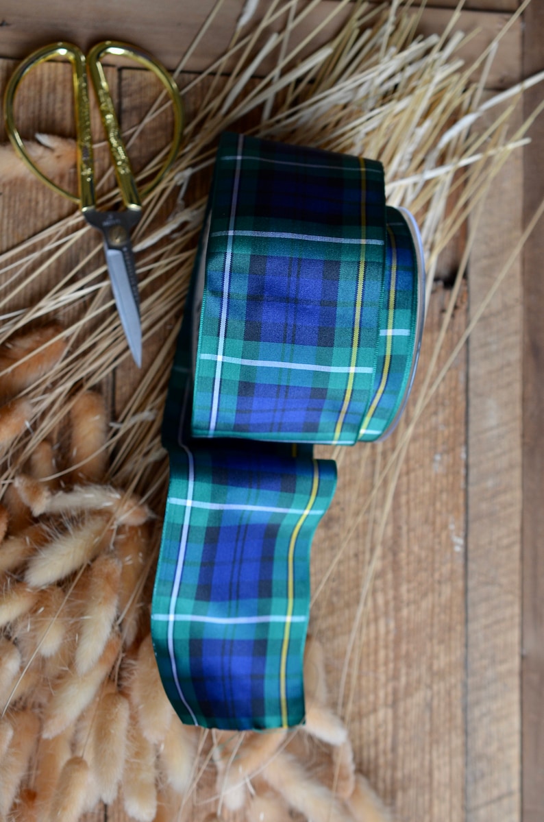 May include: A roll of blue, green, and yellow plaid ribbon with a white background. The ribbon is on a wooden surface with dried grass.