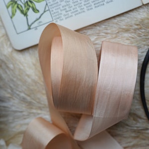 May include: A close-up of a pale peach satin ribbon, folded and twisted. The ribbon has a subtle sheen and a soft, luxurious texture.