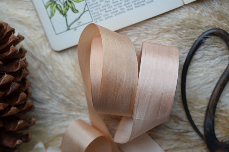 May include: A close-up of a pale peach satin ribbon, folded and twisted. The ribbon has a subtle sheen and a soft, luxurious texture.
