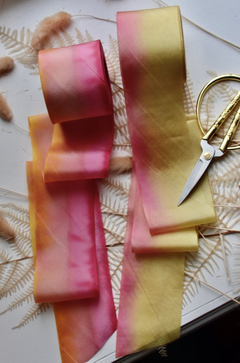May include: Two ombre silk ribbons, one in shades of pink and yellow, the other in shades of yellow and gold, are arranged on a white surface with dried fern fronds. A pair of gold scissors is also visible.