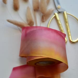 May include: A roll of ombre ribbon with a gradient of yellow, orange, and pink. The ribbon is lying on a white surface with a pair of gold scissors.