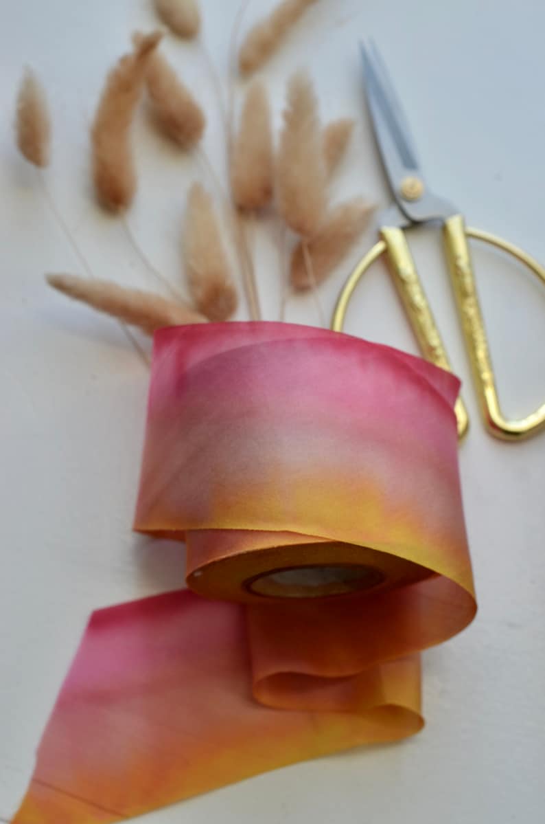 May include: A roll of ombre ribbon with a gradient of yellow, orange, and pink. The ribbon is lying on a white surface with a pair of gold scissors.