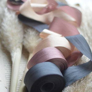 May include: A collection of four spools of ribbon in various shades of pink, beige, and black. The ribbons are arranged on a white surface with a book and dried grasses.