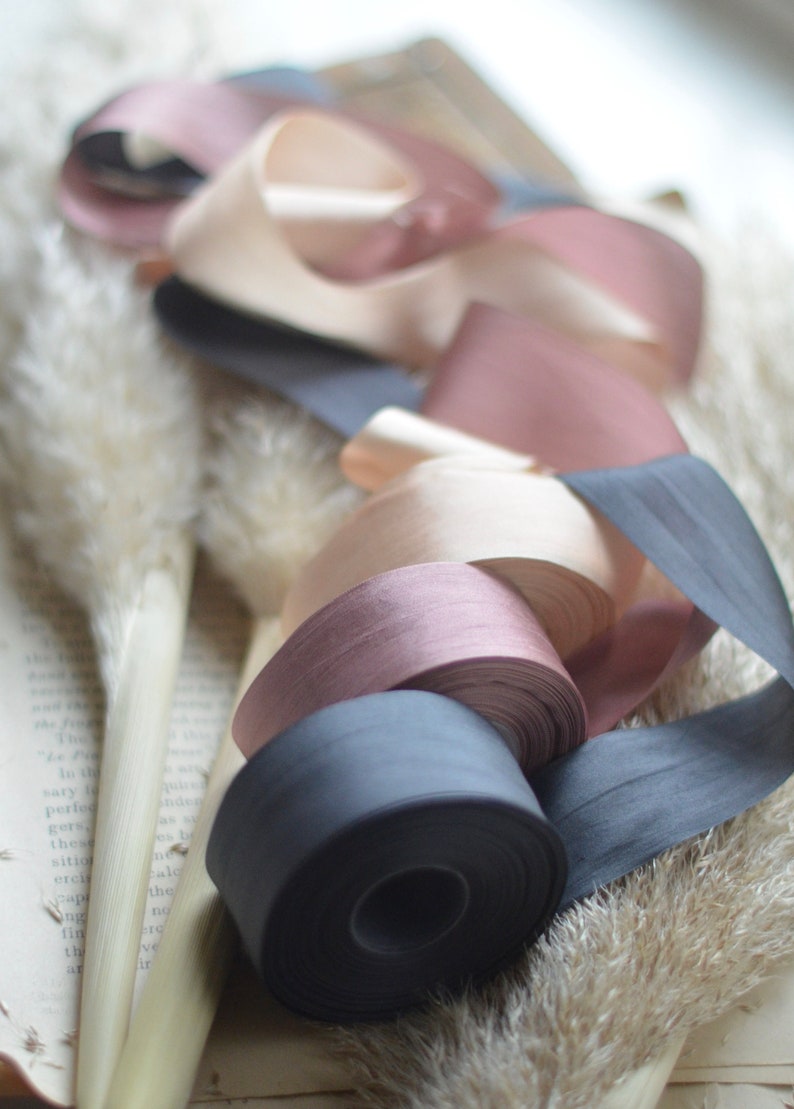 May include: A collection of four spools of ribbon in various shades of pink, beige, and black. The ribbons are arranged on a white surface with a book and dried grasses.
