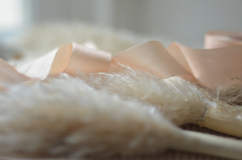 May include: A close-up of a fluffy white pampas grass bouquet tied with a soft peach satin ribbon.