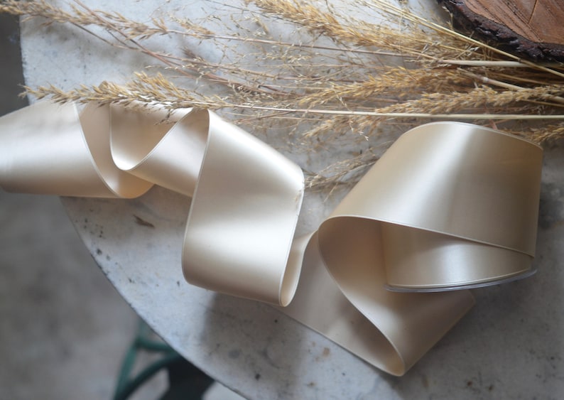 May include: A roll of ivory satin ribbon, folded and displayed on a white surface. The ribbon is smooth and shiny, with a subtle sheen.