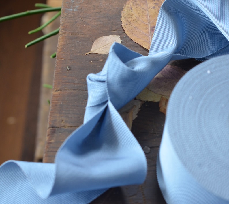 May include: A roll of light blue ribbon with a textured surface. The ribbon is folded into a bow shape and is lying on a wooden surface.