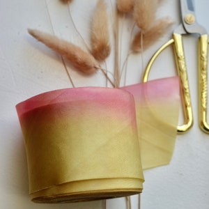 May include: A roll of ombre ribbon with a gradient of yellow to pink. The ribbon is rolled up and sitting on a white surface. There are dried pampas grasses and gold scissors in the background.