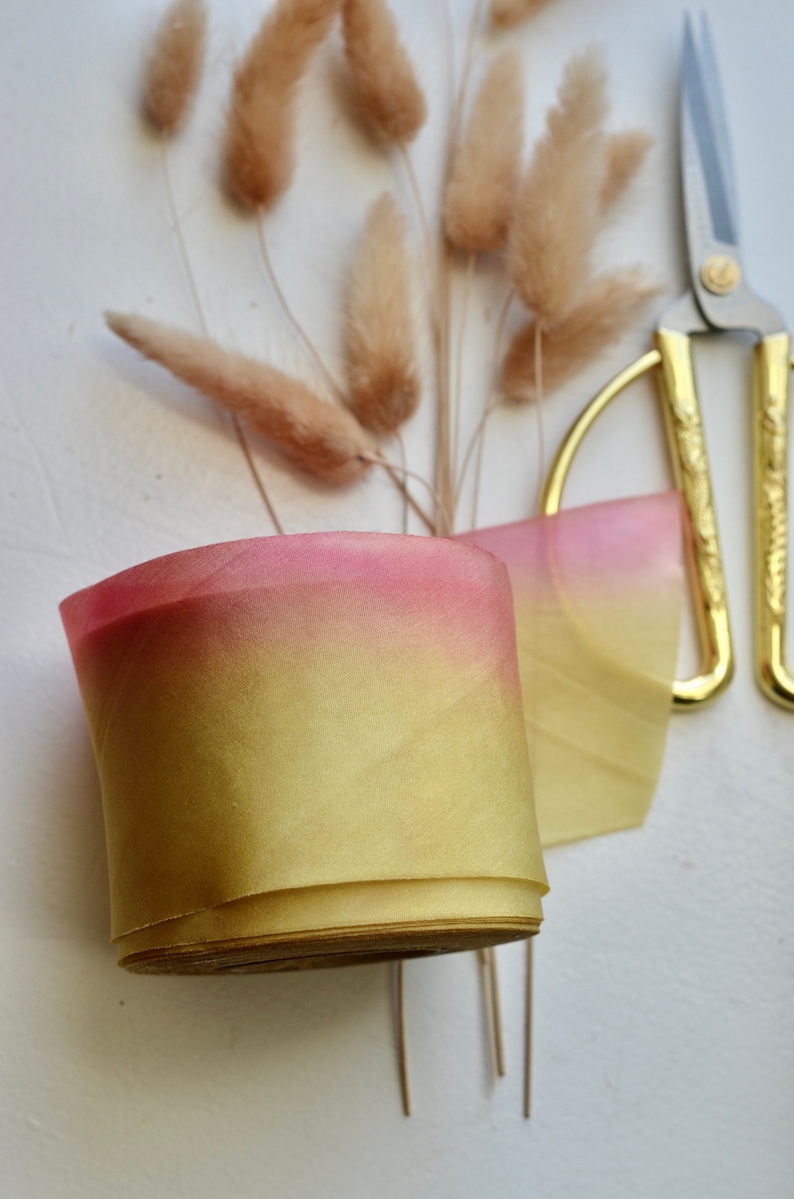 May include: A roll of ombre ribbon with a gradient of yellow to pink. The ribbon is rolled up and sitting on a white surface. There are dried pampas grasses and gold scissors in the background.