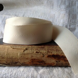 May include: A roll of cream-colored herringbone ribbon sits atop a weathered, antique book. The ribbon is neatly coiled, with a portion unraveled, displaying its textured pattern. The background is a soft, off-white fabric.