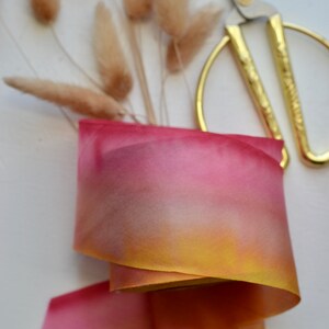 May include: A roll of ombre silk ribbon with a gradient of yellow, orange, and pink colors. The ribbon is lying on a white surface with dried pampas grass and gold scissors.