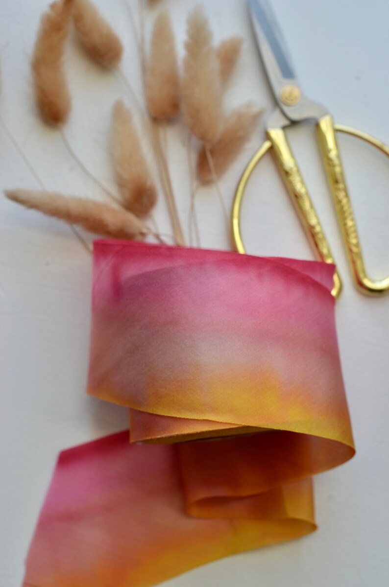 May include: A roll of ombre silk ribbon with a gradient of yellow, orange, and pink colors. The ribbon is lying on a white surface with dried pampas grass and gold scissors.