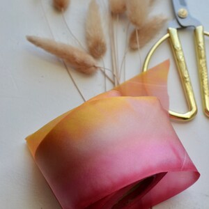 May include: A roll of ombre ribbon with a yellow to pink gradient. The ribbon is rolled up and tied with a thin white ribbon. The ribbon is on a white surface with a pair of gold scissors and a bunch of dried pampas grass.