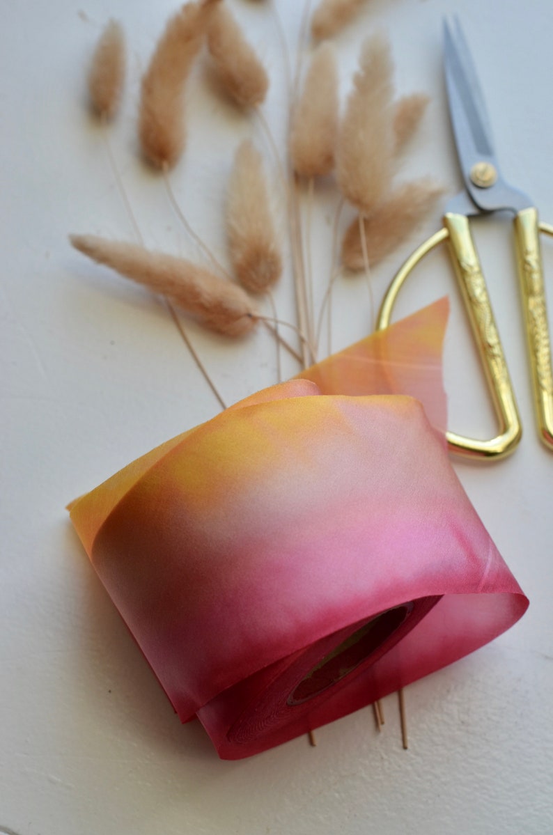 May include: A roll of ombre ribbon with a yellow to pink gradient. The ribbon is rolled up and tied with a thin white ribbon. The ribbon is on a white surface with a pair of gold scissors and a bunch of dried pampas grass.