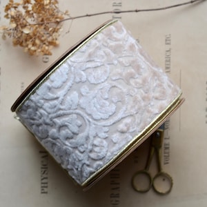 May include: A roll of cream-colored velvet ribbon with a floral pattern, edged with gold trim. The ribbon is brown on the sides. The background is a vintage book page with text.