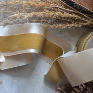 May include: A roll of ivory satin ribbon with a gold velvet trim. The ribbon is folded and arranged on a white surface.