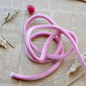 May include: Pink velvet ribbon, a soft and delicate material, is coiled and arranged on a textured surface. The ribbon is a light pink colour and has a subtle sheen.