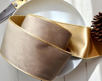 May include: A rolled-up, wide ribbon in shades of beige and gold, with gold trim, rests on a white plate. A pine cone and scissors are also visible. The ribbon is likely for crafts or gift wrapping.
