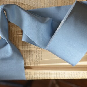May include: A light blue satin ribbon, folded and displayed on an old book. The ribbon is smooth and has a subtle sheen.