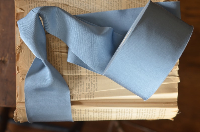 May include: A light blue satin ribbon, folded and displayed on an old book. The ribbon is smooth and has a subtle sheen.