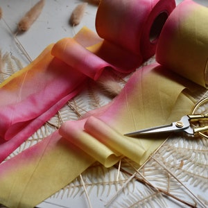 May include: Two rolls of ombre ribbon in shades of yellow and pink, with one roll partially unrolled. A pair of gold scissors are lying on a white surface with dried fern leaves.