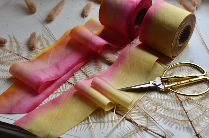 May include: Two rolls of ombre ribbon in shades of yellow and pink, with one roll partially unrolled. A pair of gold scissors are lying on a white surface with dried fern leaves.