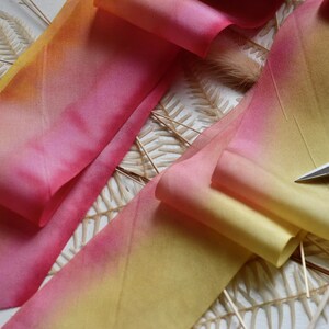 May include: A close-up of a yellow and pink ombre ribbon with a pair of silver scissors on top. The ribbon is folded and arranged on a white surface with dried fern branches.