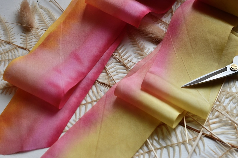 May include: A close-up of a yellow and pink ombre ribbon with a pair of silver scissors on top. The ribbon is folded and arranged on a white surface with dried fern branches.