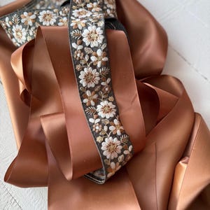 May include: A close-up of a brown satin ribbon with a floral embroidered trim. The trim features white and gold flowers on a dark gray background.