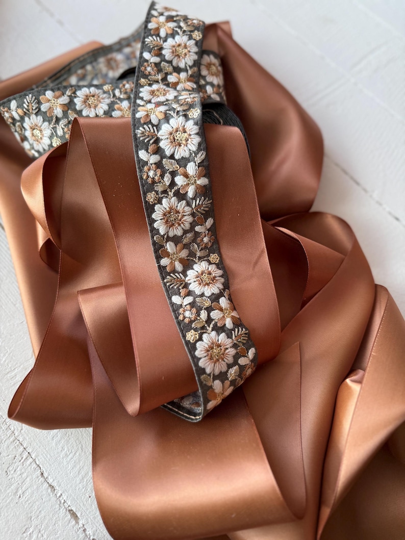 May include: A close-up of a brown satin ribbon with a floral embroidered trim. The trim features white and gold flowers on a dark gray background.