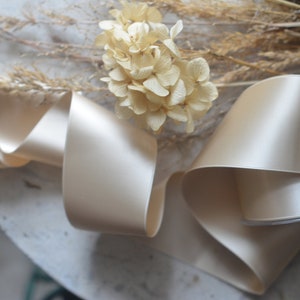 May include: A close-up of a beige satin ribbon, folded and arranged on a white surface. The ribbon is smooth and shiny, with a subtle sheen. Dried flowers are visible in the background.