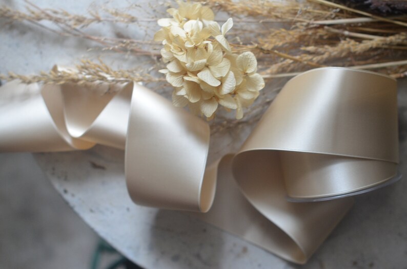 May include: A close-up of a beige satin ribbon, folded and arranged on a white surface. The ribbon is smooth and shiny, with a subtle sheen. Dried flowers are visible in the background.