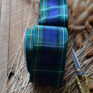 May include: A roll of green, blue, and yellow plaid ribbon with a pair of gold scissors on a wooden surface.