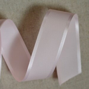 May include: A pale pink satin ribbon with a ribbed texture. The ribbon is curled and folded, showing the smooth satin edge.