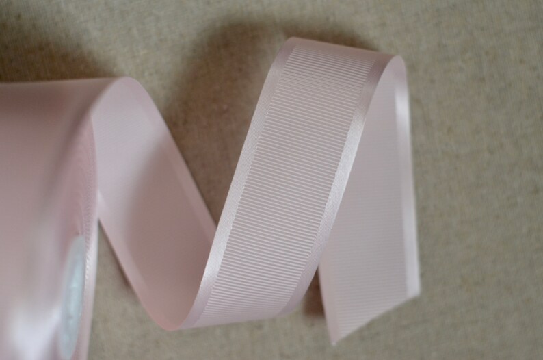 May include: A pale pink satin ribbon with a ribbed texture. The ribbon is curled and folded, showing the smooth satin edge.