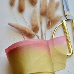 May include: A roll of ombre ribbon with a pink to yellow gradient. The ribbon is wrapped around a cardboard spool. The ribbon is next to a pair of gold scissors and a bunch of dried pampas grass.