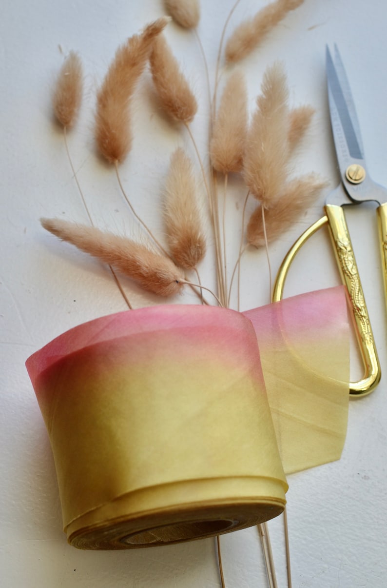 May include: A roll of ombre ribbon with a pink to yellow gradient. The ribbon is wrapped around a cardboard spool. The ribbon is next to a pair of gold scissors and a bunch of dried pampas grass.