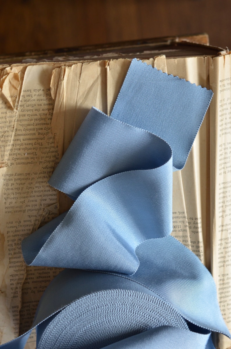 May include: A roll of light blue grosgrain ribbon with a textured surface. The ribbon is folded and arranged on top of a vintage book.