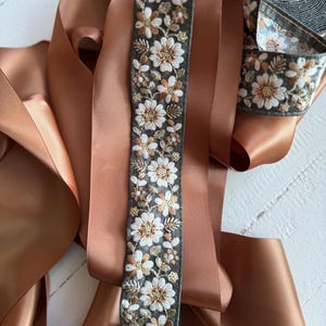 May include: A roll of brown satin ribbon with a floral embroidered trim. The trim features white flowers with gold accents on a dark green background.
